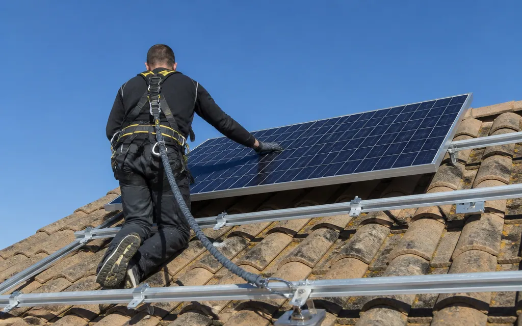 Installateur harnaché fixant un panneau solaire sur une toiture en pente, crochets posés sur la tuile